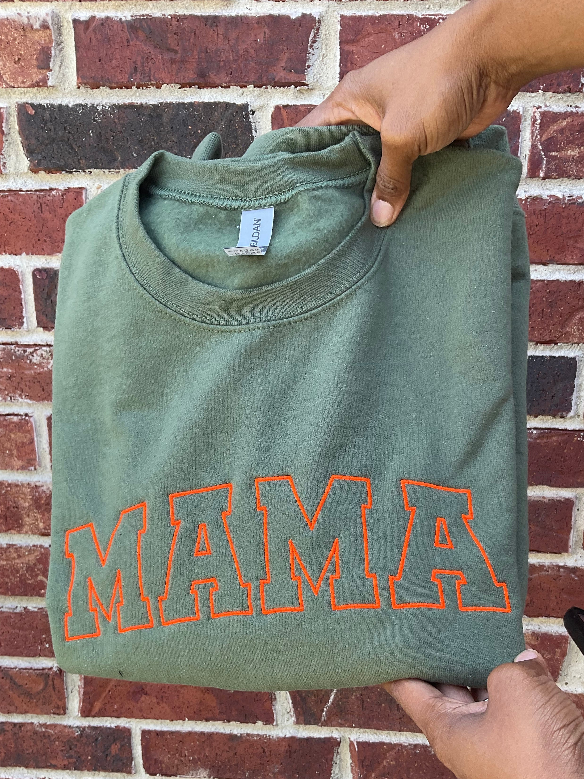 Folded green t-shirt with 'MAMA' printed in orange on a brick background